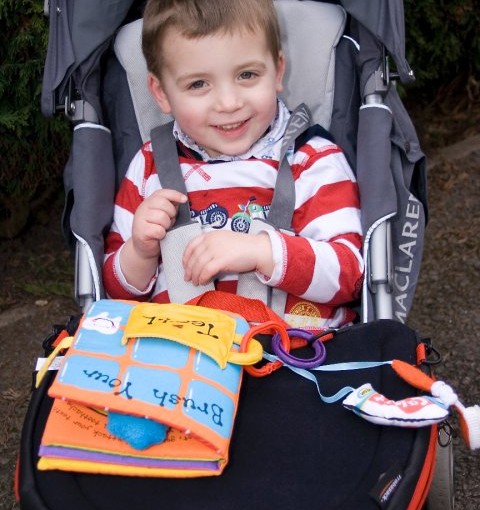Victor sits in his buggy, smiling to the camera, with a Trabasack on his lap with books and toys attached to it