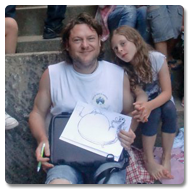 Brian sits outdoors on stone steps, smiling to the camera, he is surrounded by lots of local children whilst he illustrates drawings on using his Trabasack lap desk