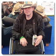 Mark is sitting in a wheelchair with a wry expression, he is wearing a Cowboy-style straw hat, placing a can of beer on the tray surface of the Trabasack Mini on his lap