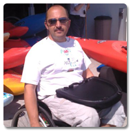 Alan looks straight to the camera, he is sitting in a wheelchair with a Trabasack Curve on his lap, behind him are canoes lined-up ready to be used