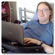 Danni is lying in bed, propped-up with cushions, she smiles to the camera whilst using her laptop on the Trabasack lap desk