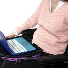 Image is a photograph of a lady wearing a peach-coloured, ribbed jumper, sitting in a wheelchair with a purple trim Trabasack Curve on her lap, using a laptop on the desk surface