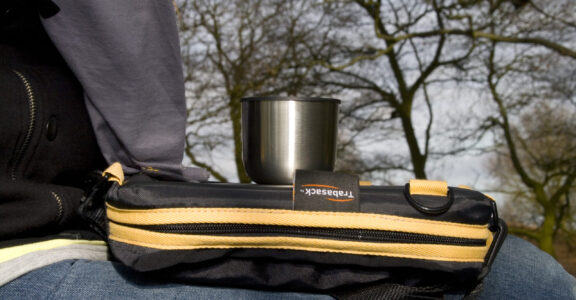Camping and Picnic Tray Photo of Trabasack Mini on someone's lap with a cup on top, behind in the background is a forrest of trees