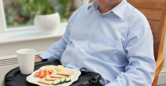 An older man sits in a high-back chair, eating lunch using the Trabasack Curve as a lap tray