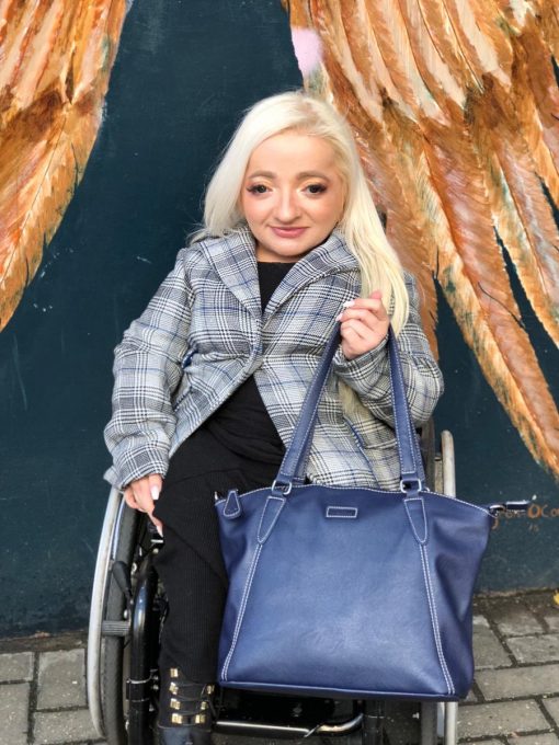 Photograph shows Sam Renke dressed in a plaid jacket and black trousers, seated in a wheelchair holding a Navy blue Samantha bag in her left hand.