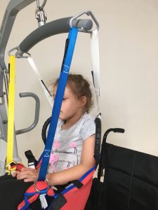 Image shows a photograph of the side view of a young girl sat in the ProMove transfer sling attached to a spreader bar hoist