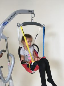 Image shows a photograph of a young girl being cradled in a ProMove Sling attached to a spreader bar hoist