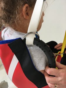 Image shows a photograph of the back, rear shoulder view of a young girl sitting in a ProMove sling with the hoist straps being attached