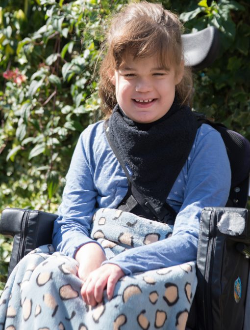 Image shows a photograph of a smiling young girl with brown hair, sat outside in a wheelchair, wearing a black towelling kerchief around her neck with a blue and beige leopard print fleece blanket covering her knees