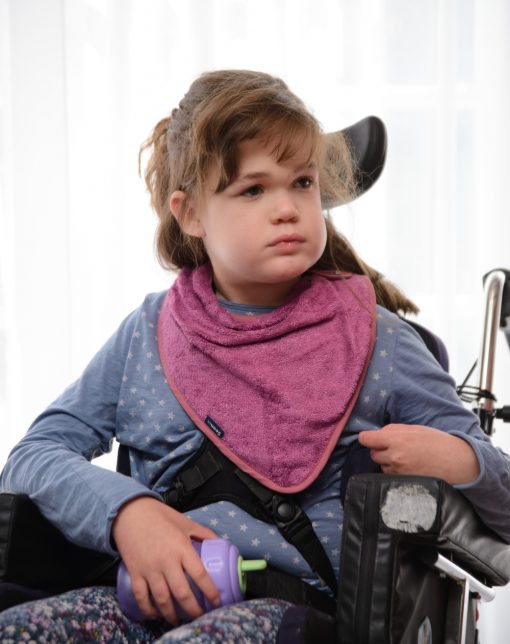Image shows a photograph of a young girl sat in a wheelchair, wearing pretty floral leggins and a pink Seenin bamboo side fastening kerchief