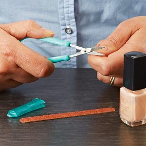 Image is a photograph of someone using the mini easi-grip scissors to trim their nails, with a bottle of nail varnish and a nail file on the table in front of them.