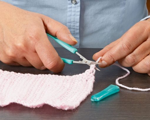 Image is a photograph of someone using the mini easi-grip scissors to cut-off a loose end from a piece of knitted fabric.