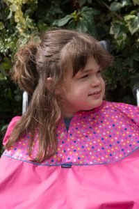 Image is a photograph of a young girl with brown-hair smiling and looking away from the camera, sat outside in a wheelchair wearing a Pink Spot design waterproof total wheelchair cover in bright pink, with blue, purple and yellow spot-design