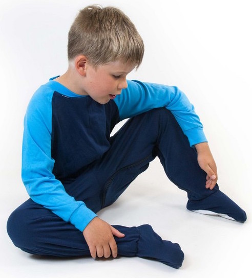 Image shows a photograph of a boy seated casually on a white background, wearing a footed sleepsuit in navy and turquoise