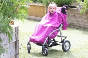 Image is a photograph of a girl smiling at the camera, whilst sat in an SEN stroller whilst covered with a fuchsia pink total fleece cover