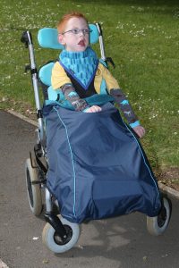Image shows a photograph of a boy in an adaptive SEN stroller, sitting outdoors wearing a blue wheelchair leg protector