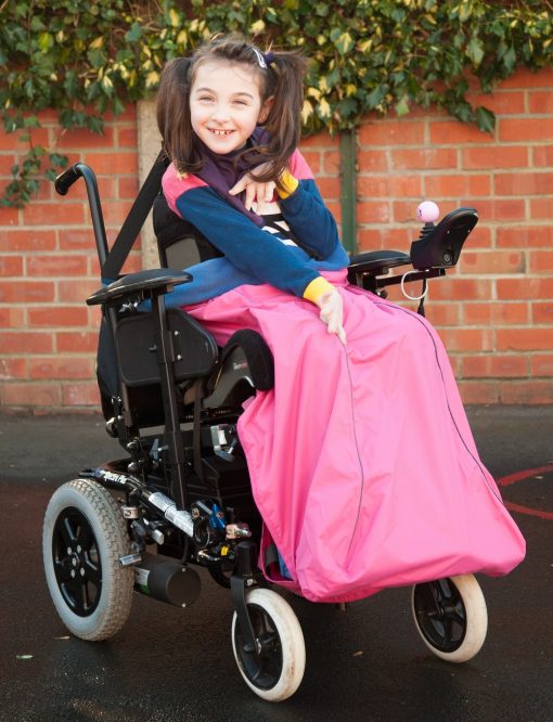 Image is a photograph of a cheerful girl with brown hair in bunches, sat outdoors in a wheelchair wearing a pink wheelchair leg cover