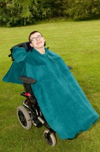 Image is a photograph of a young man smiling, sitting in a wheelchair outdoors with a teal full cover fleece