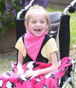 Image shows a photograph of a laughing young girl, sat outside in a wheelchair, wearing a bright pink kerchief around her neck with a pink sheep-patterned fleece blanket upon her knees.