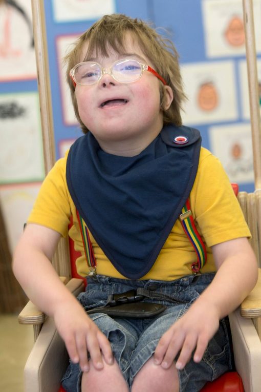 Image shows a photograph of a cheerful young boy wearing brightly coloured trouser braces and t-shirt, and a navy blue kerchief around his neck