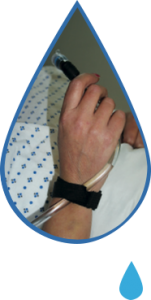 Image is a photograph cut-out in a tear-drop shape, showing a close-up of a patient wearing a hospital gown, with the drinking tube of the Water Drop attached to the side of their hand using a velcro strap, accessing water to drink