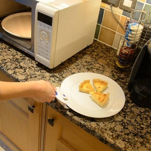 Image is a photograph of a kitchen counter top showing a plate of quiche being removed from a microwave using a Buckingham Coolhand