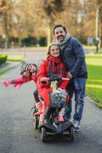 Image is a photgraph of a family of mum, dad and daughter outdoors with mum sat on a mobility scooter, dad standing close and smiling, and daughter sat behind on a Skoe Hitch with her head titled smiling at the camera.