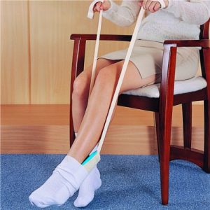 Image is a photograph of the lowerhalf of a woman seated in a dining chair using the Deluxe Sock Aid to put-on a pair of white socks