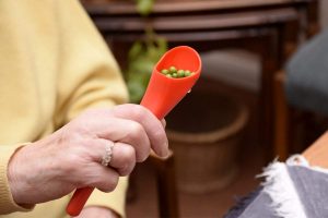 Image is a photograph of an older lady wearing a yellow jumper, holding a S'Up Spoon Mini filled with garden peas