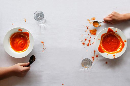 Image is a photograph of two bowls of tomato soup at either end of a clothed table. Next to the first bowl sits a S'Up Spoon with relatively little mess to the table cloth. Next to the second bowl sits a traditional soup spoon, with tomato soup spillage on the table cloth