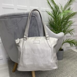 Image is a photograph of a Matt Silver Samantha Renke accessible handbag in the extra size, hanging by the straps over the back of a grey velvet chair in a living room.