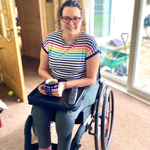 Image is a photograph of Elizabeth Gwilliam wearing a colourful stripy top, in a wheelchair using the Trabasack Mini as a wheelchair tray for her cup of tea and remote control